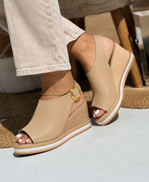 Primer plano de modelo luciendo sandalias de plataforma tipo cuña en color taupe con diseño cerrado en el talón, correa ajustable con hebilla dorada y suela cómoda de doble tono.