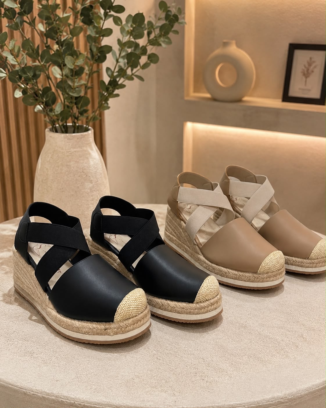 sandalias de plataforma tipo espadrille, un par en negro y otro en taupe, con capellada de cuero, bandas elásticas cruzadas en el empeine y suela cubierta de yute, exhibidas en un estante.